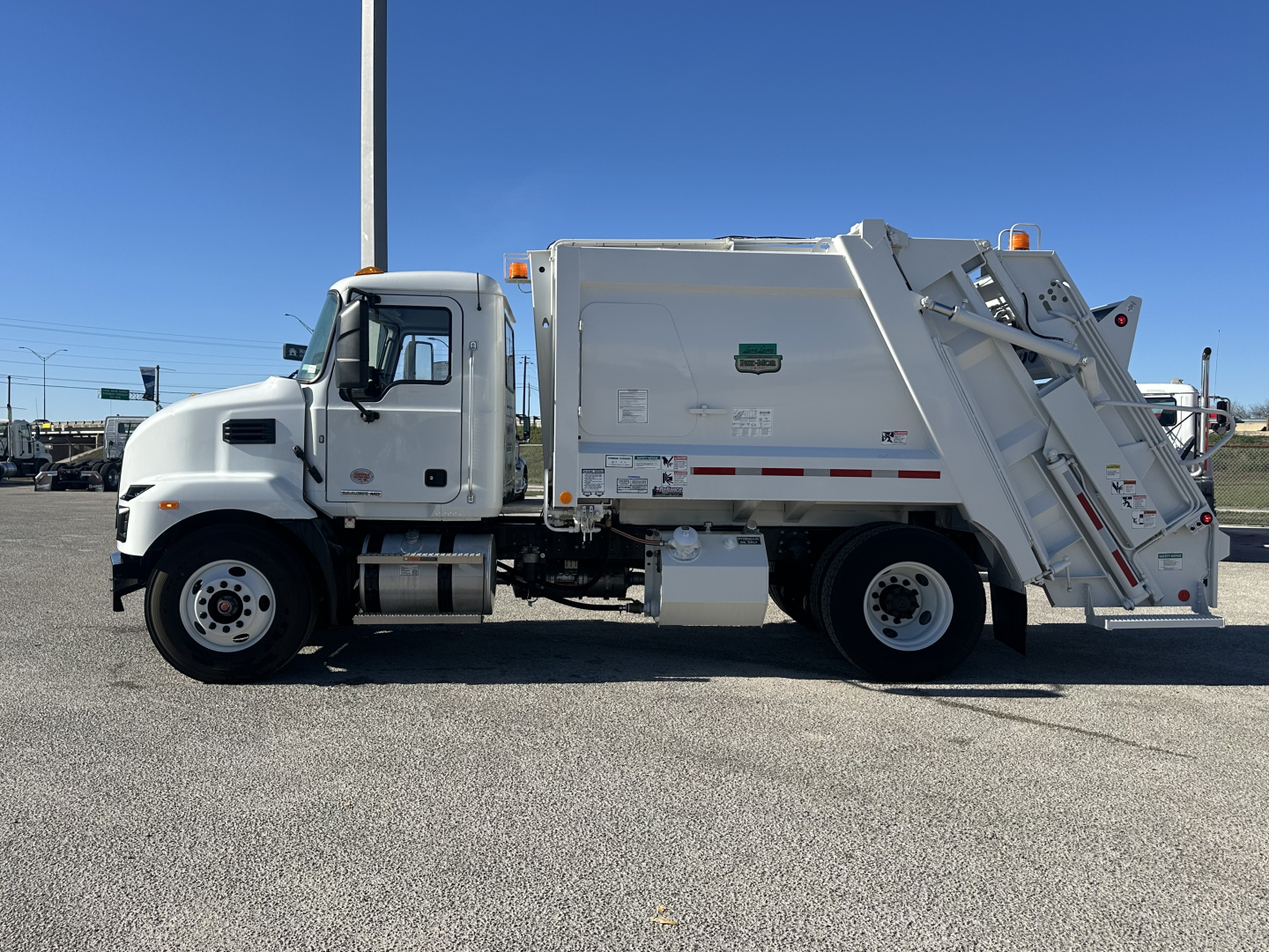 Truck side view of a white Mack MD6 refuse truck showing cab and rear compactor body with warning decals and reflective tape.