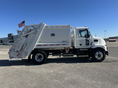 Mack MD6 rear-load refuse truck in side profile view with hydraulic compactor and rear hopper, white cab and body, American flag in background.
