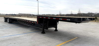 Flatbed trailer showing a long black drop deck with landing gear and spread legs in a parking lot.