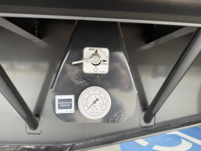 Trailer display panel showing a gauge and controls: a small lever with a label, a round pressure gauge, and a suspension pressure sticker. Visible text includes 'RAISE' and 'LOWER' around the lever, a circular gauge with numbers, and a smal