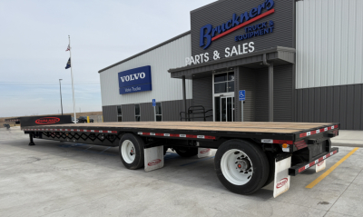 Flatbed trailer with wooden deck and black frame in front of a truck equipment building.