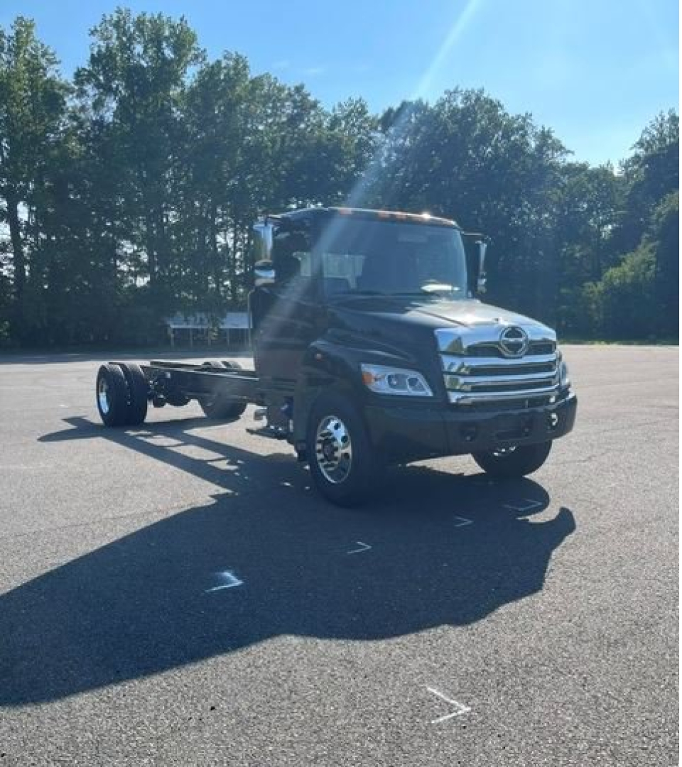 Truck cab chassis: forward view of a new Hino L7 cab chassis in a parking lot, black exterior, chassis behind cab, chrome grille, dual rear wheels, daytime sun