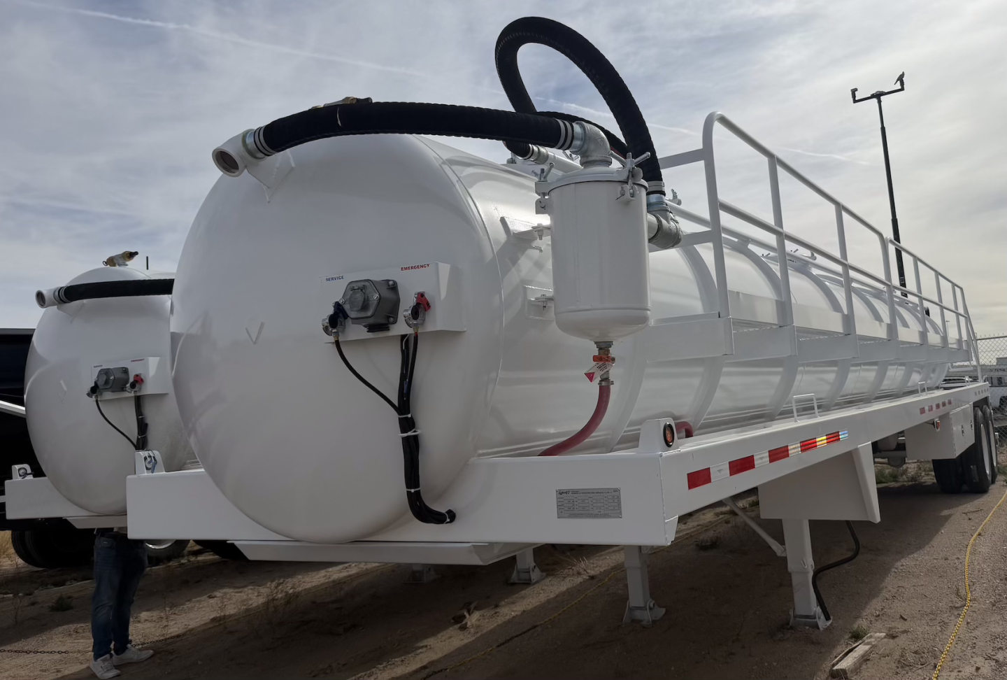 Vacuum trailer tank showing large cylindrical white steel tank with inlet hoses and manway, on a low-bed trailer chassis.