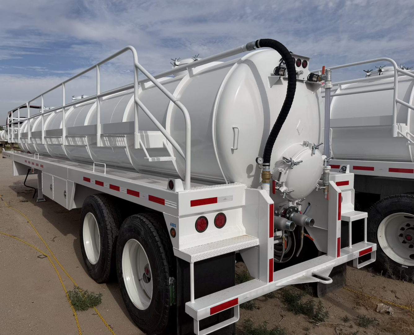 Flatbed trailer with a large white vacuum steel tank, side rails, and rear equipment. Text not clearly legible; visible features include circular manhole and hoses.