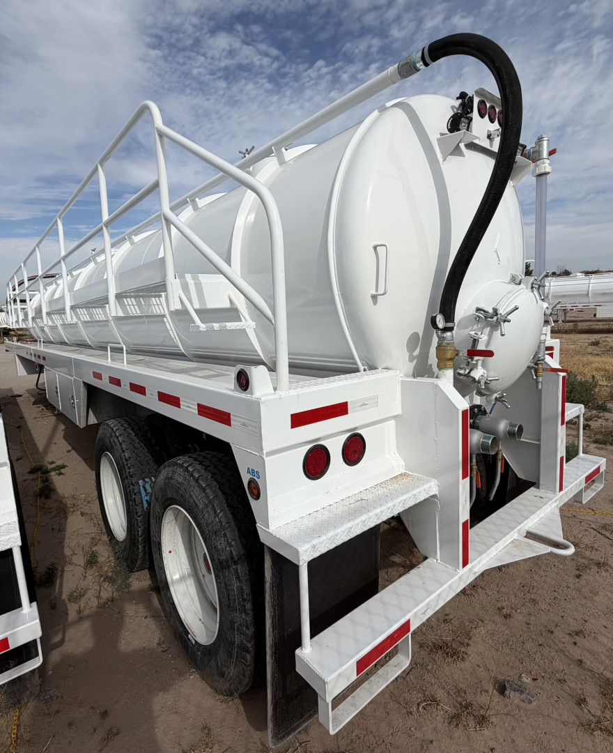 Vacuum tanker trailer with white cylindrical tank on a tri-axle chassis, rear manway, hoses, and railing; rear access steps and taillights visible.