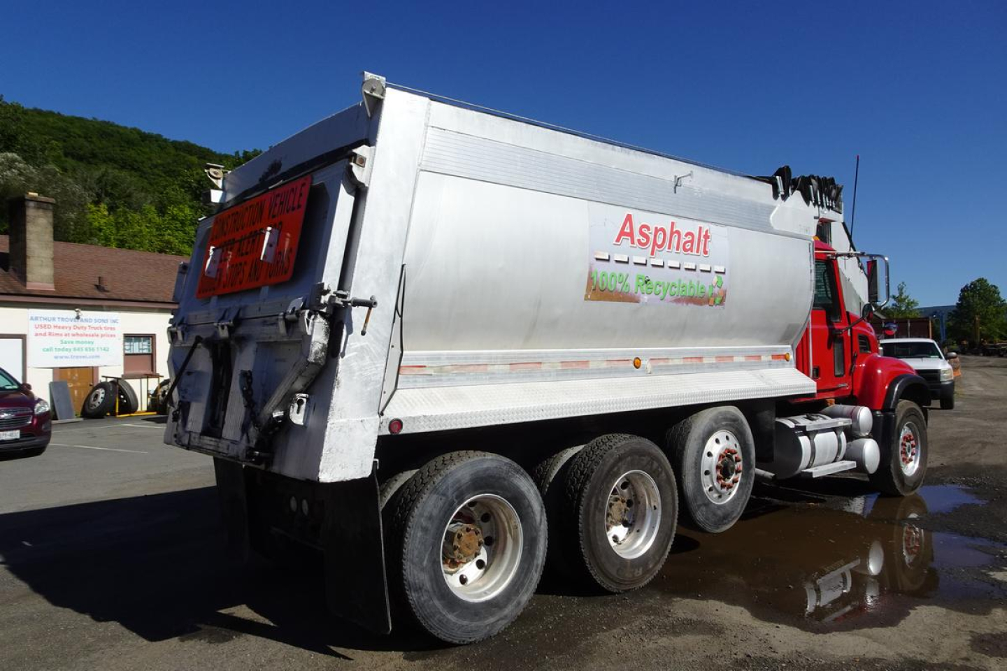 Dump truck with silver open-top dump body showing Asphalt recycling logo on side; red cab; three rear axles; Mack CV Granite CV713 style; visible text includes 'Asphalt', '100% Recycled' and caution signs on rear.