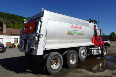 Dump truck with silver open-top dump body showing Asphalt recycling logo on side; red cab; three rear axles; Mack CV Granite CV713 style; visible text includes 'Asphalt', '100% Recycled' and caution signs on rear.