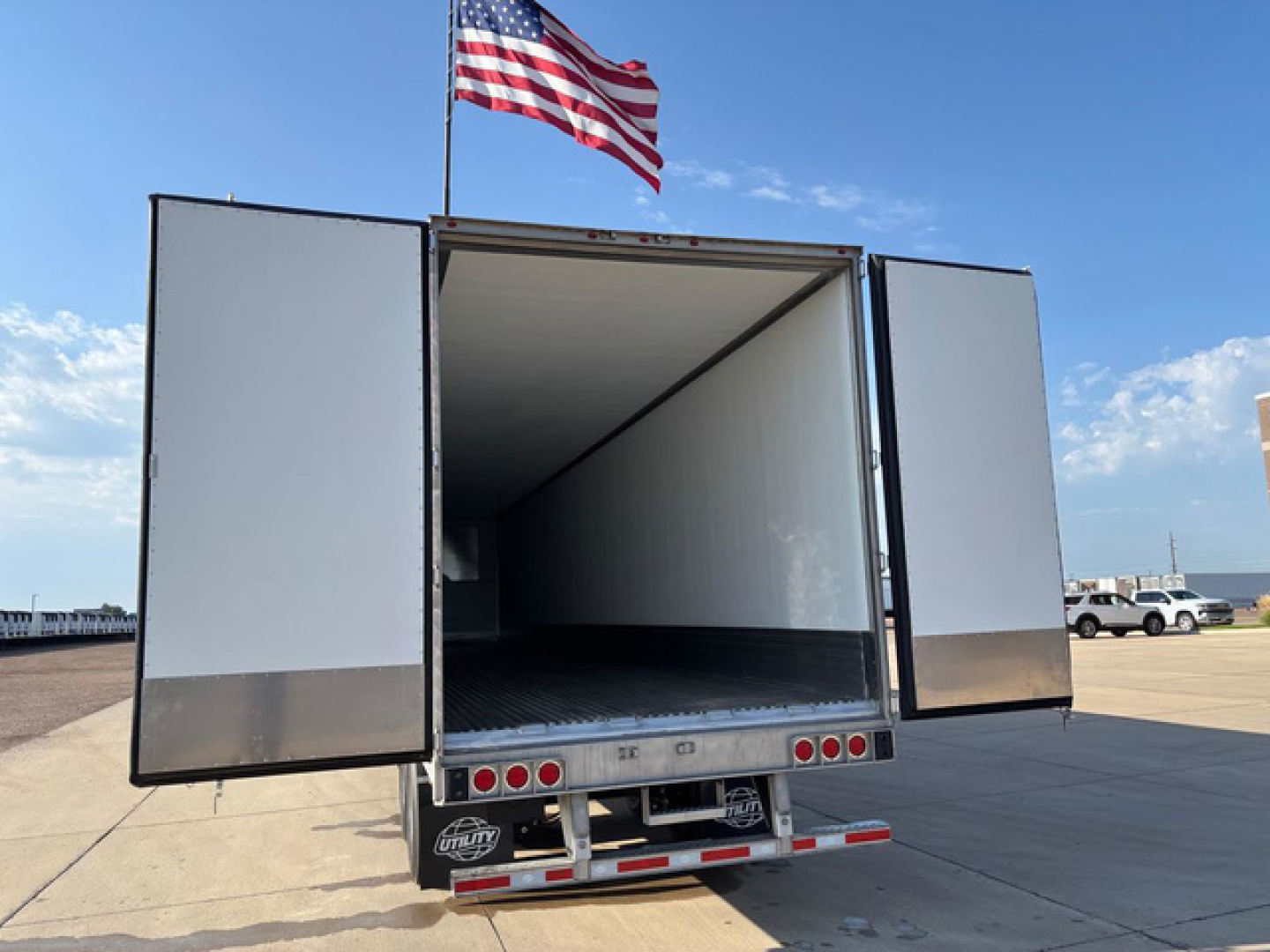 Utility reefer trailer with open rear doors showing empty cargo area and metal flooring; American flag flying above, clear blue sky.