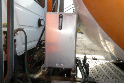 Boxed electrical controller mounted on chassis with cables and metal platform; partial view of a white truck cab on left and an orange-tinted tank or shield on right.