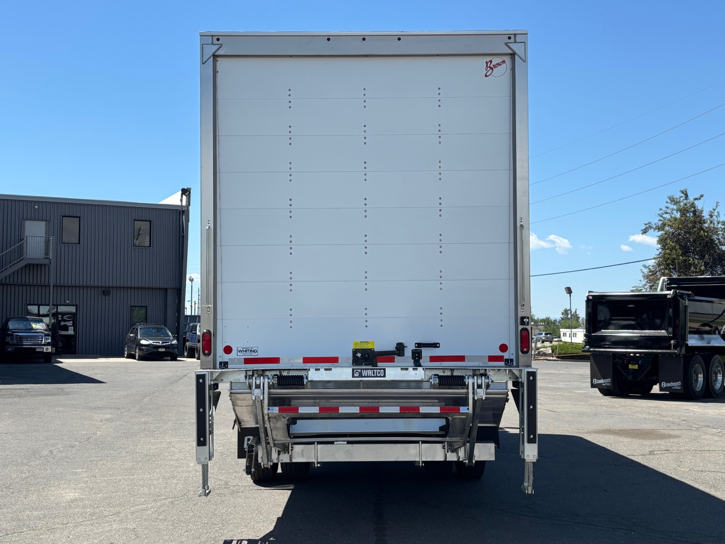 Rear view of a new 2026 Mack MD6 box truck with closed cargo area, rear underride guard, and stabilizing legs extended. VIN tag not visible.