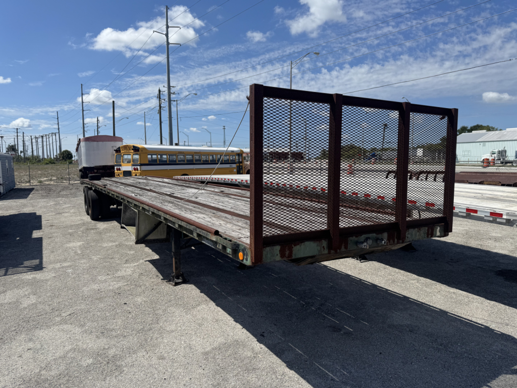 Flatbed trailer with a metal ramp gate attached, long wooden deck, and a yellow school bus in the background.
