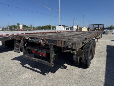 Flatbed trailer showing weathered wooden deck and rear with three red tail lights, dual wheels, and rusted bumper. Open lot with other trailers in background.