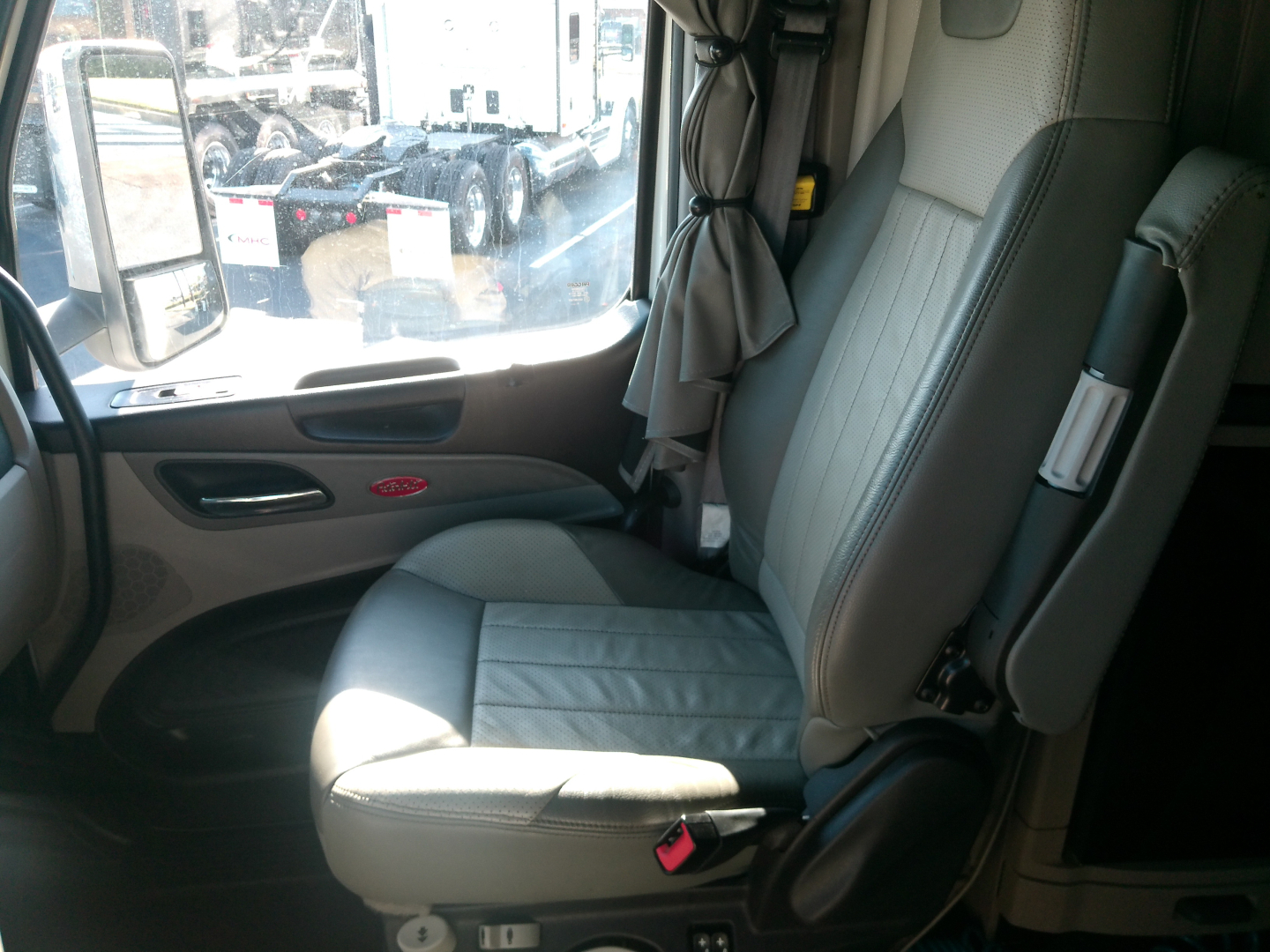 Peterbilt truck cab interior showing driver seat with light gray upholstery, window and door panel visible, curtain in the background.