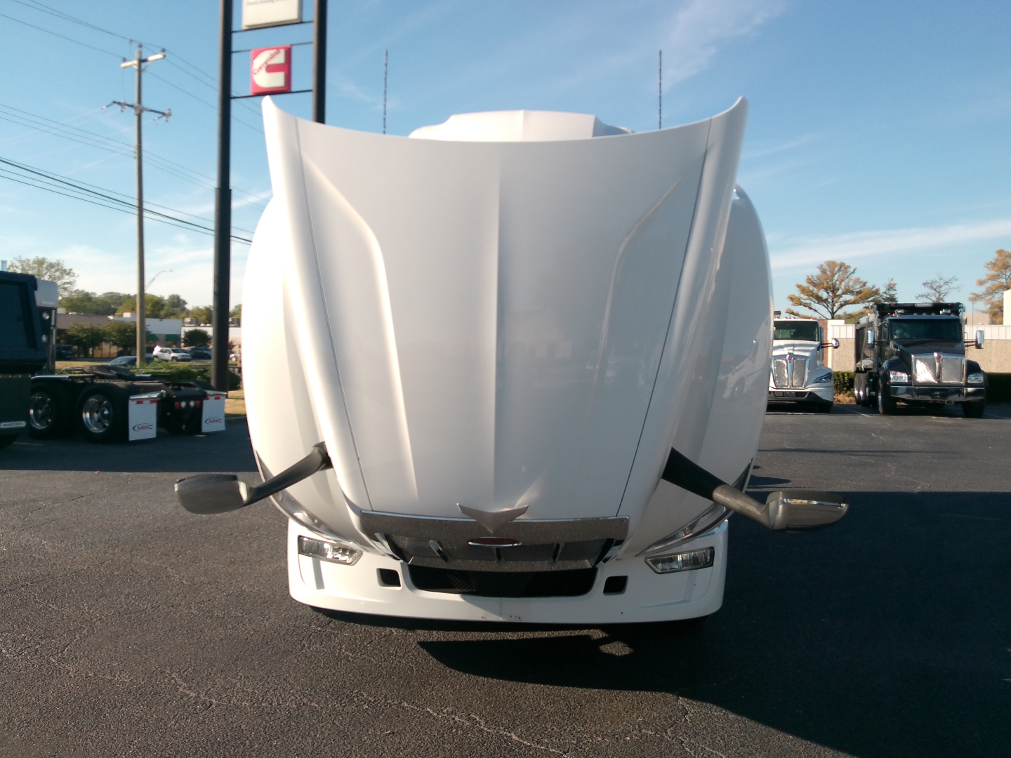 Peterbilt 579 front view with hood raised on the truck in a dealership lot. The image shows the front bumper, headlights, and emblem on the grille; other trucks visible in background.