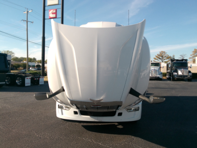 Peterbilt 579 front view with hood raised on the truck in a dealership lot. The image shows the front bumper, headlights, and emblem on the grille; other trucks visible in background.