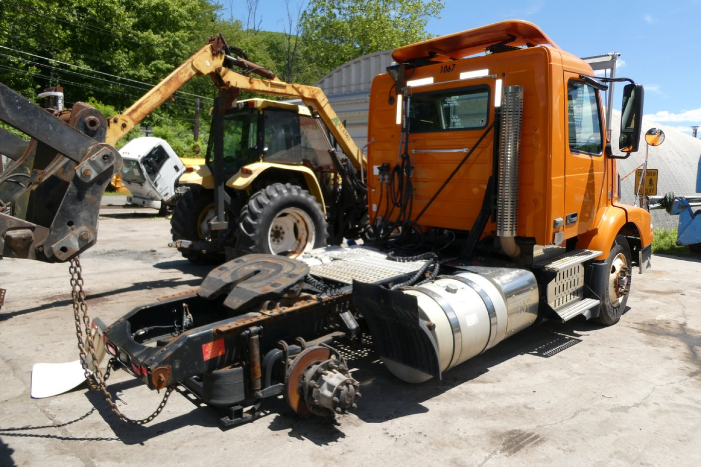 Orange Volvo VNM42T daycab truck with front sleeper cab and chassis visible, attached to a hitch and towing apparatus in a yard.