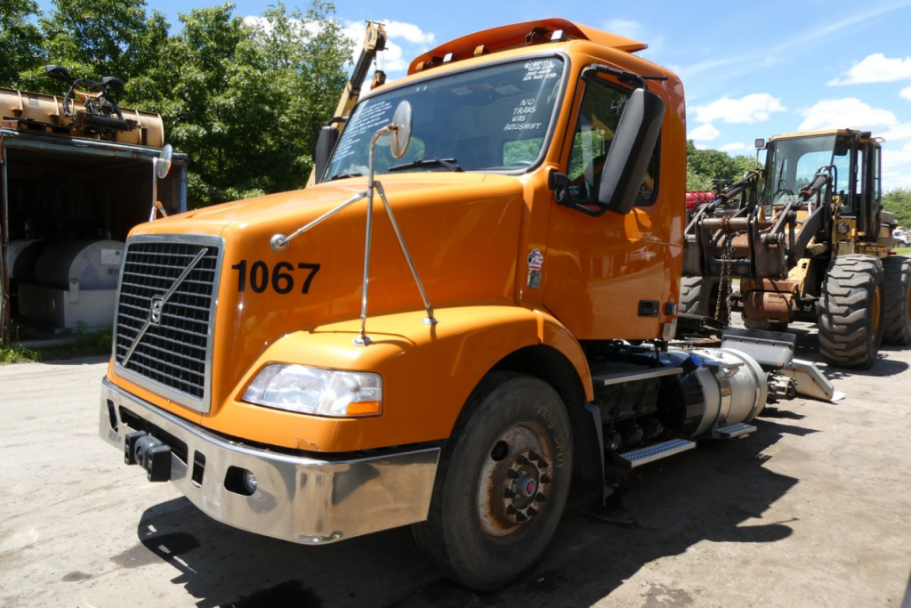 Volvo VNM42T conventional daycab truck with orange cab and black grille showing number 1067 on door.