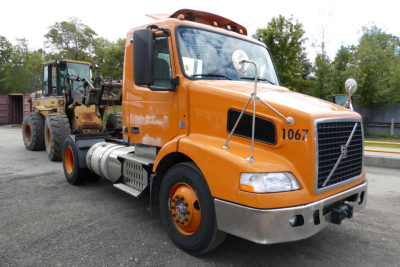Volvo VNM42T conventional daycab truck with orange cab, front grille, chrome bumper, and attached heavy equipment carrier; number 1067 on door; VIN and stock details not visible.