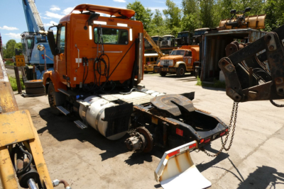 Orange Volvo VNM42T truck chassis with cab, fuel tank, and exposed wiring in a yard; surrounding equipment and trucks visible.