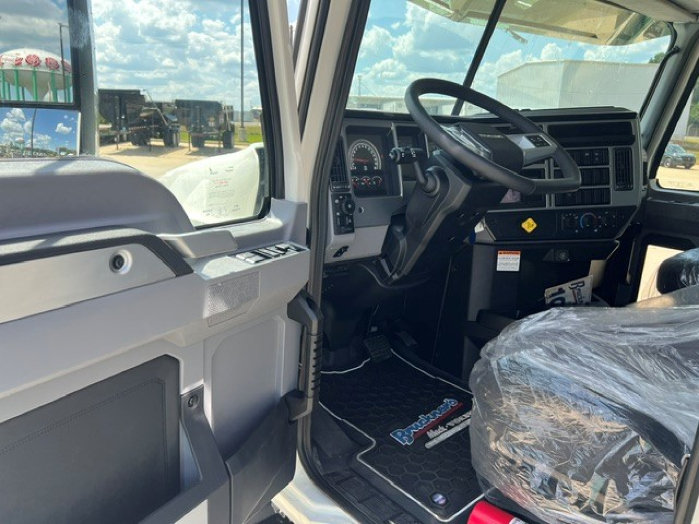 Truck cab interior showing steering wheel, dashboard, and driver door; plastic seat cover on passenger seat; floor mats with company branding; dashboard controls and instrument cluster visible.