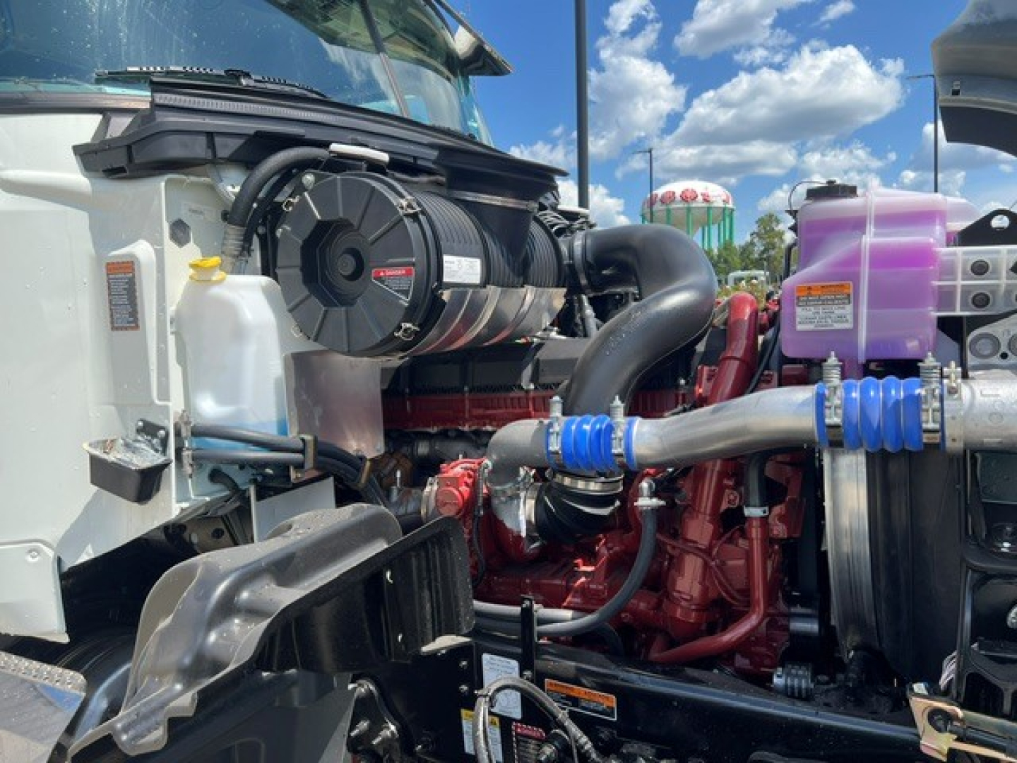 Engine bay of a 2027 Mack GR104F dump truck showing air intake, coolant reservoir, hoses, and camouflage pink S-barrel fuel lines.