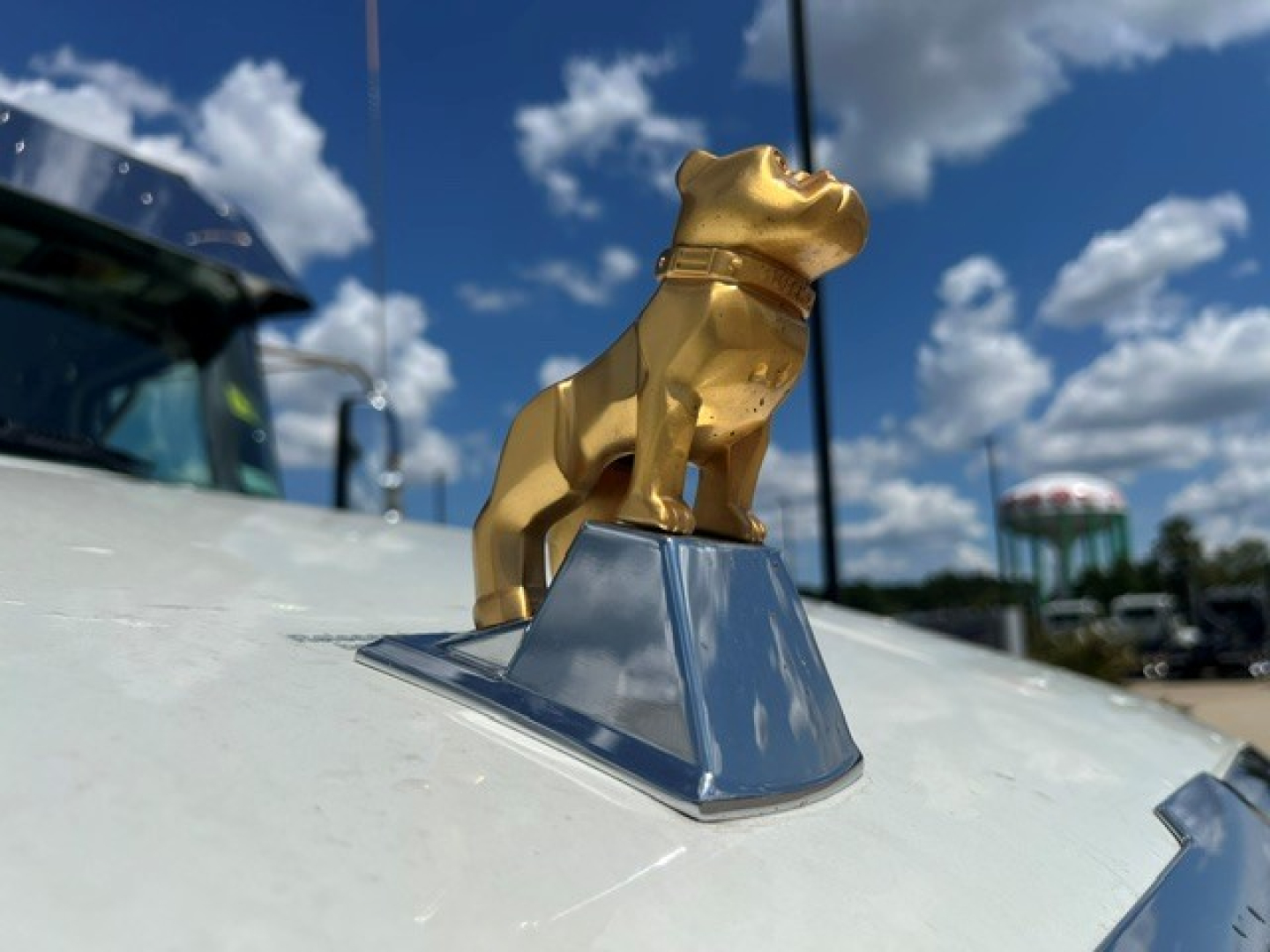 Front hood ornament of a Mack truck with a golden bulldog mascot on a chrome base, against a blue sky with clouds.