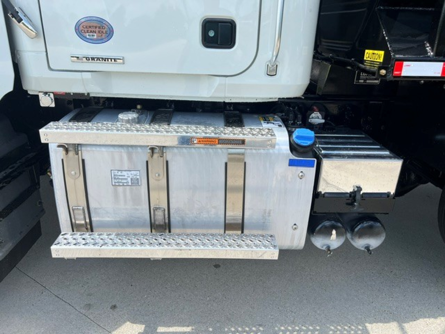 Aluminum side toolbox and fuel tanks under the cab of a white Mack GR104F dump truck showing metal steps and storage compartments.
