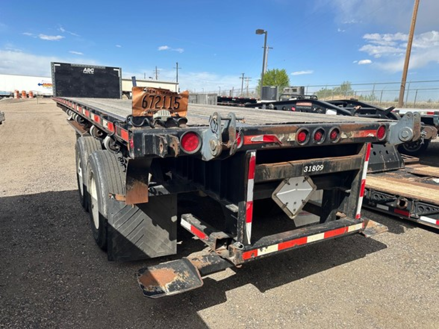 Flatbed trailer rear view with dual rear axles, black frame, red and white reflective tape, wooden deck at top, license plate area, and a metal support plate.