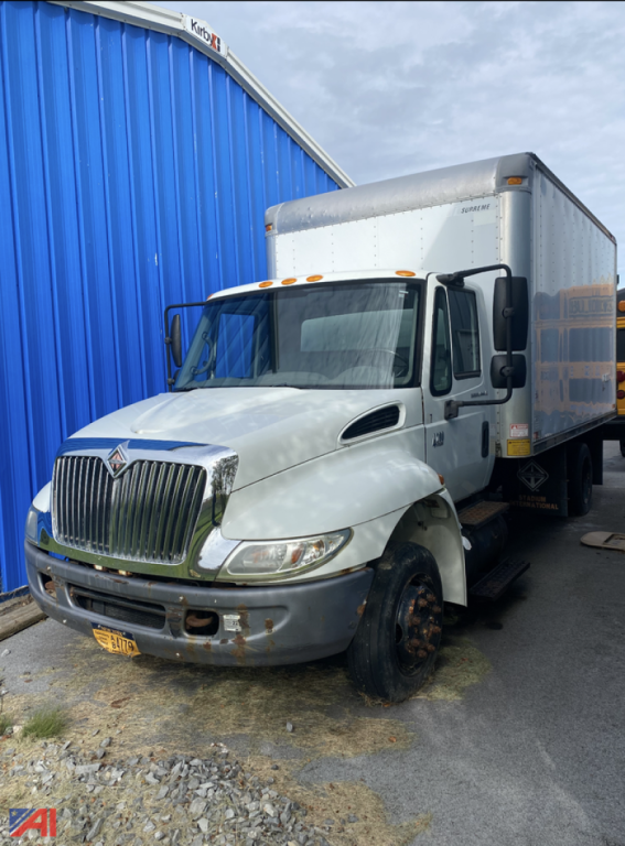 Box truck front-left view with white cab and gray box, International emblem grille, missing front wheel on the left, dirt around wheel, yellow license plate.