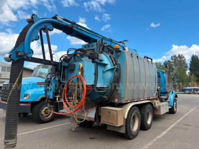 Vacuum truck with blue cab and large cylindrical tank, rear hose reel arm extended, multiple hoses coiled, on a paved lot under blue sky.