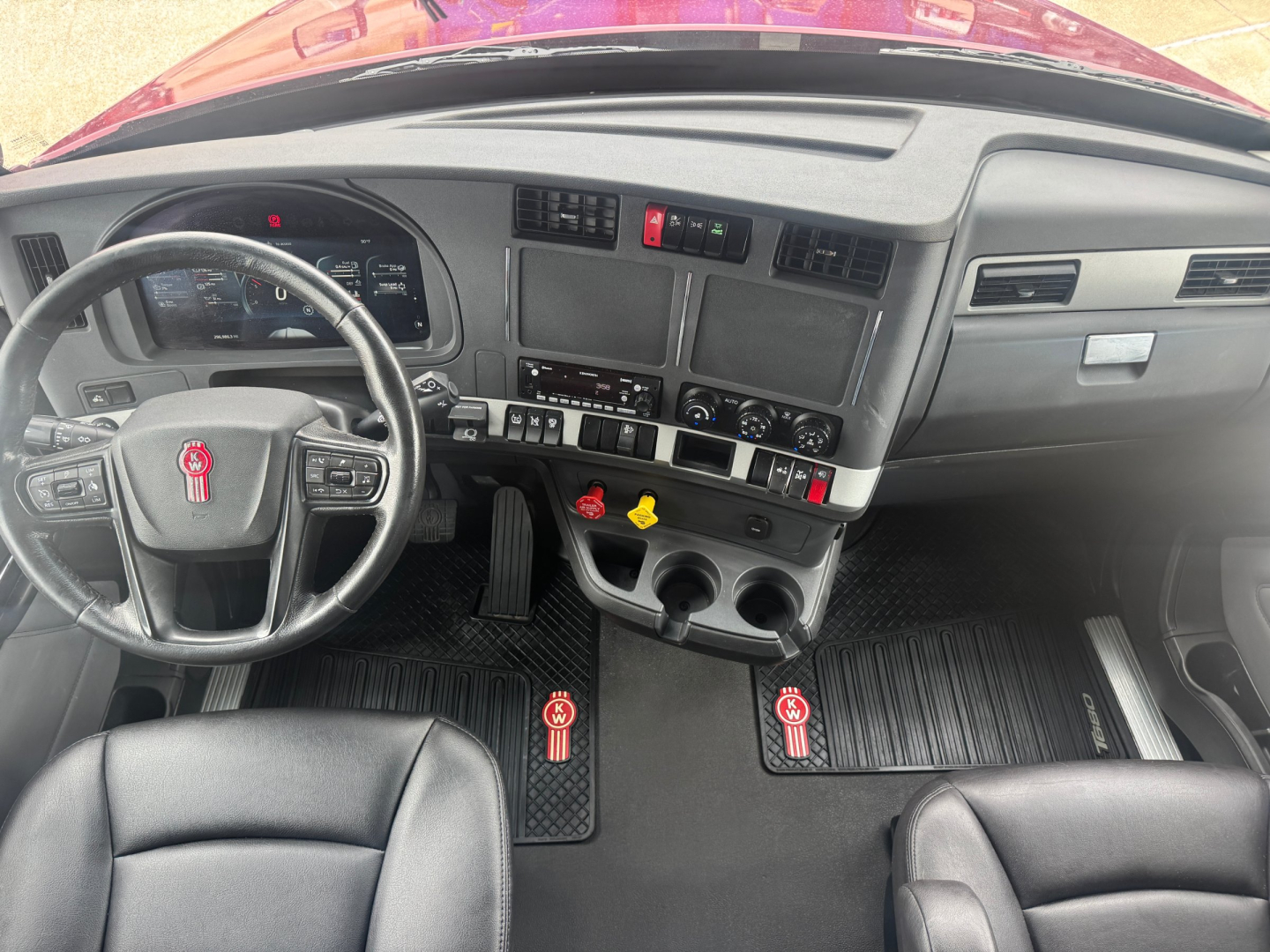 Kenworth T680 cab interior showing steering wheel, dash, center console, and floor mats with two pedals visible.