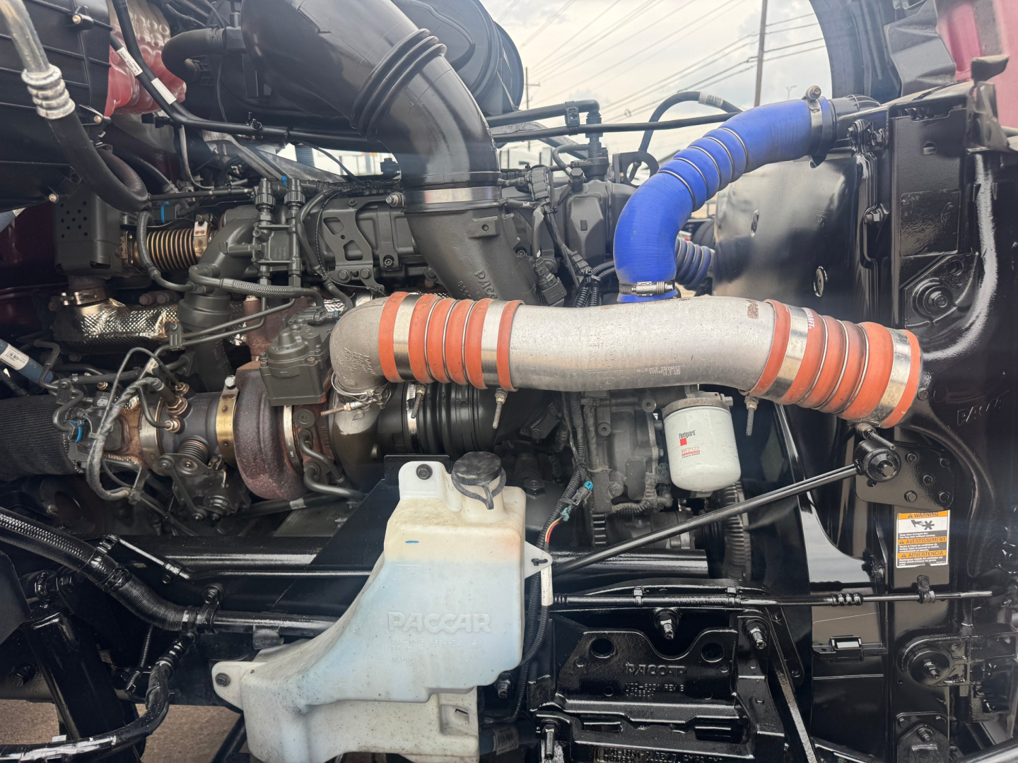 Kenworth T680 engine bay showing large turbo pipe with orange heat wrap and blue silicone hose, coolant reservoir labeled TracAir, various hoses and components.