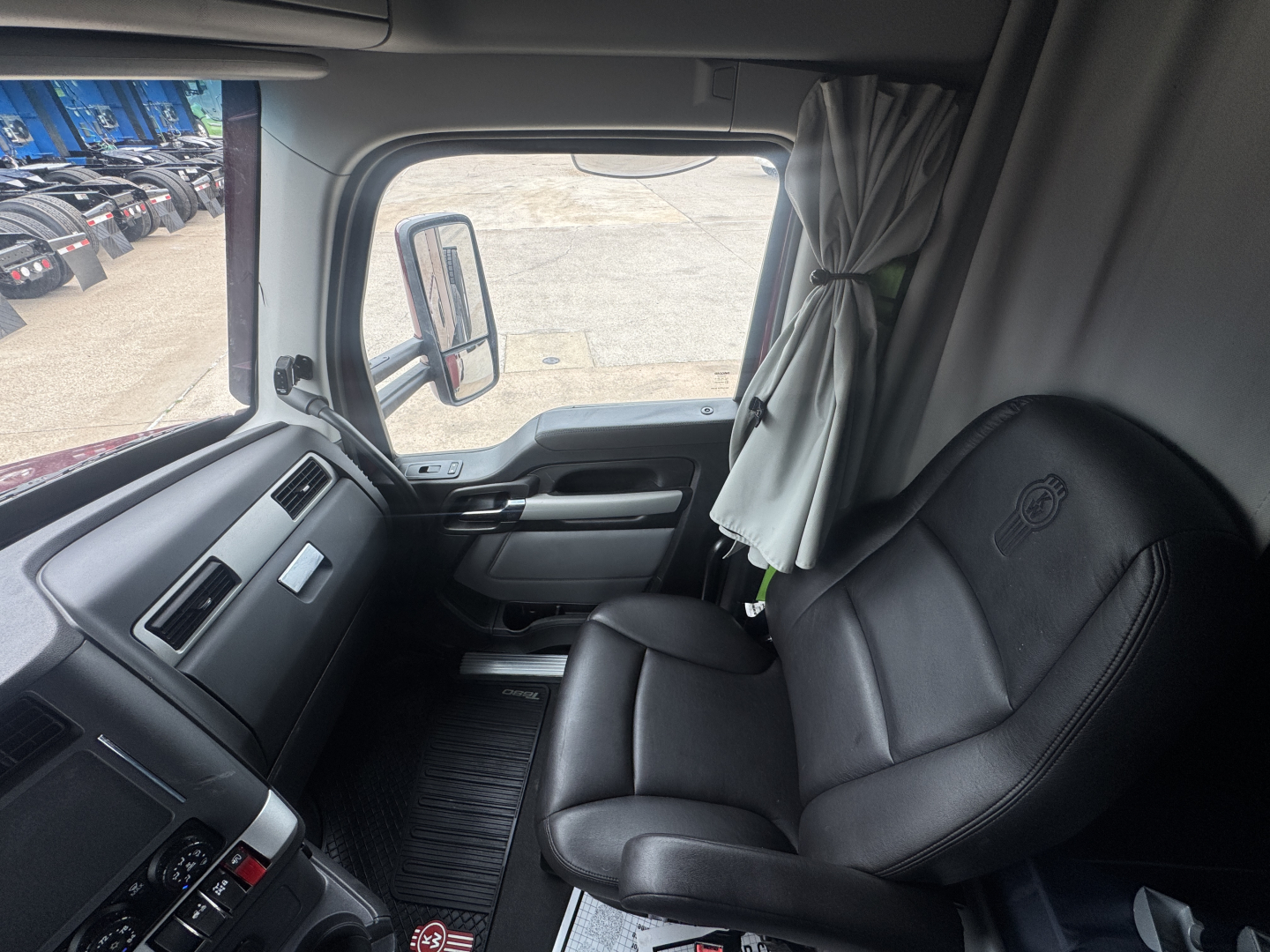 Kenworth T680 truck interior cab seat and dashboard area visible from doorway; black leather driver seat, curtain, side mirror, and window.