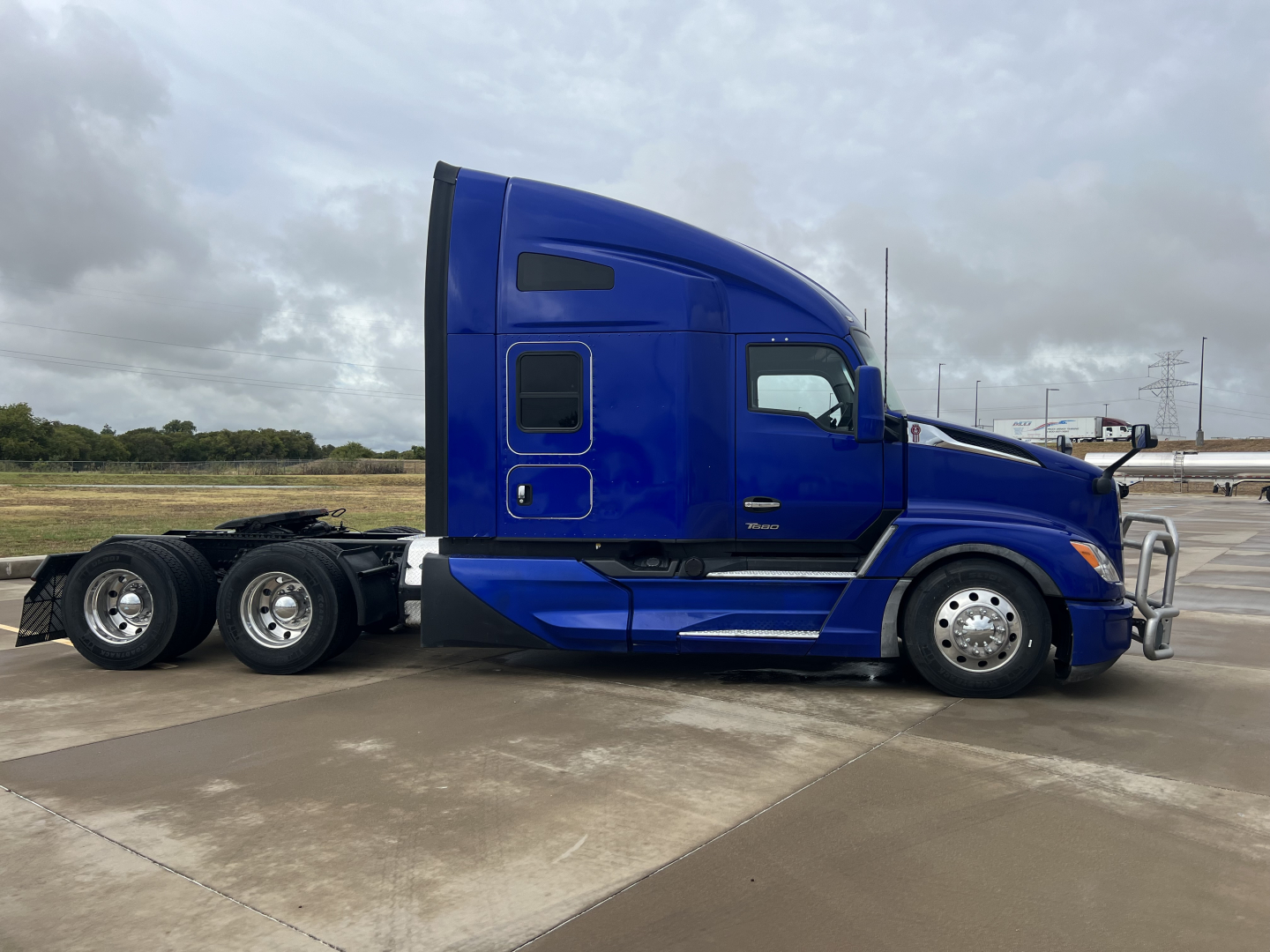 Blue Kenworth T680 tractor unit side view showing sleeper cab, chrome bumper, dual rear axles, and windows.