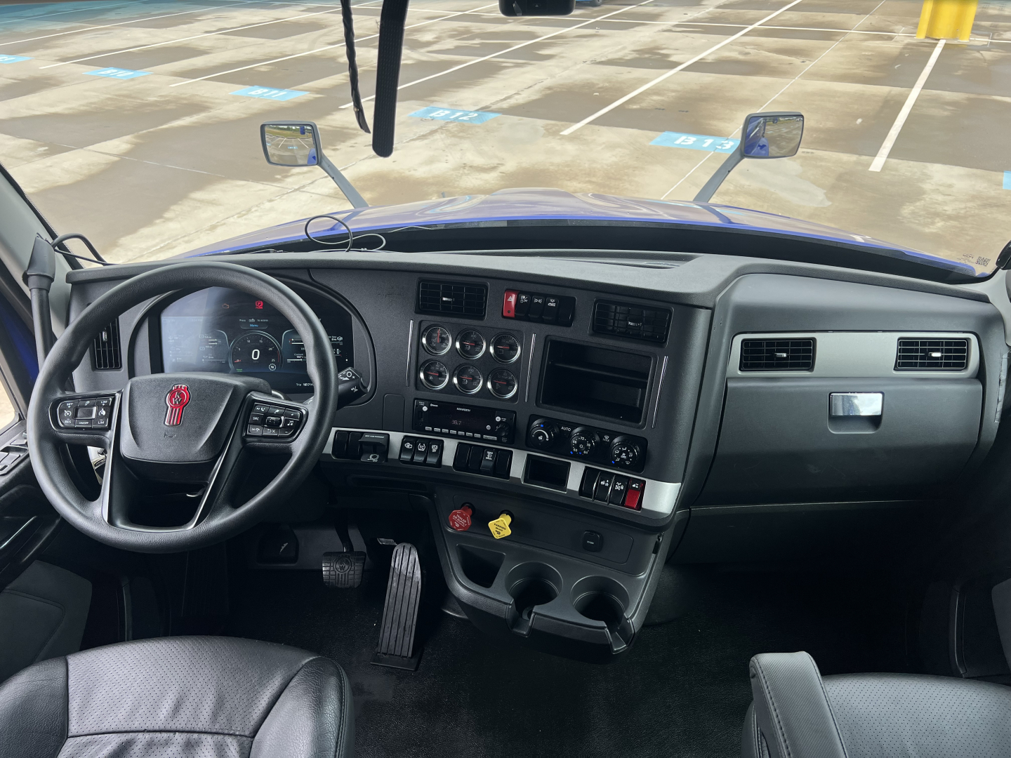 Kenworth T680 cab interior showing dashboard controls and steering wheel with tires and parking lot reflection visible through windshield.