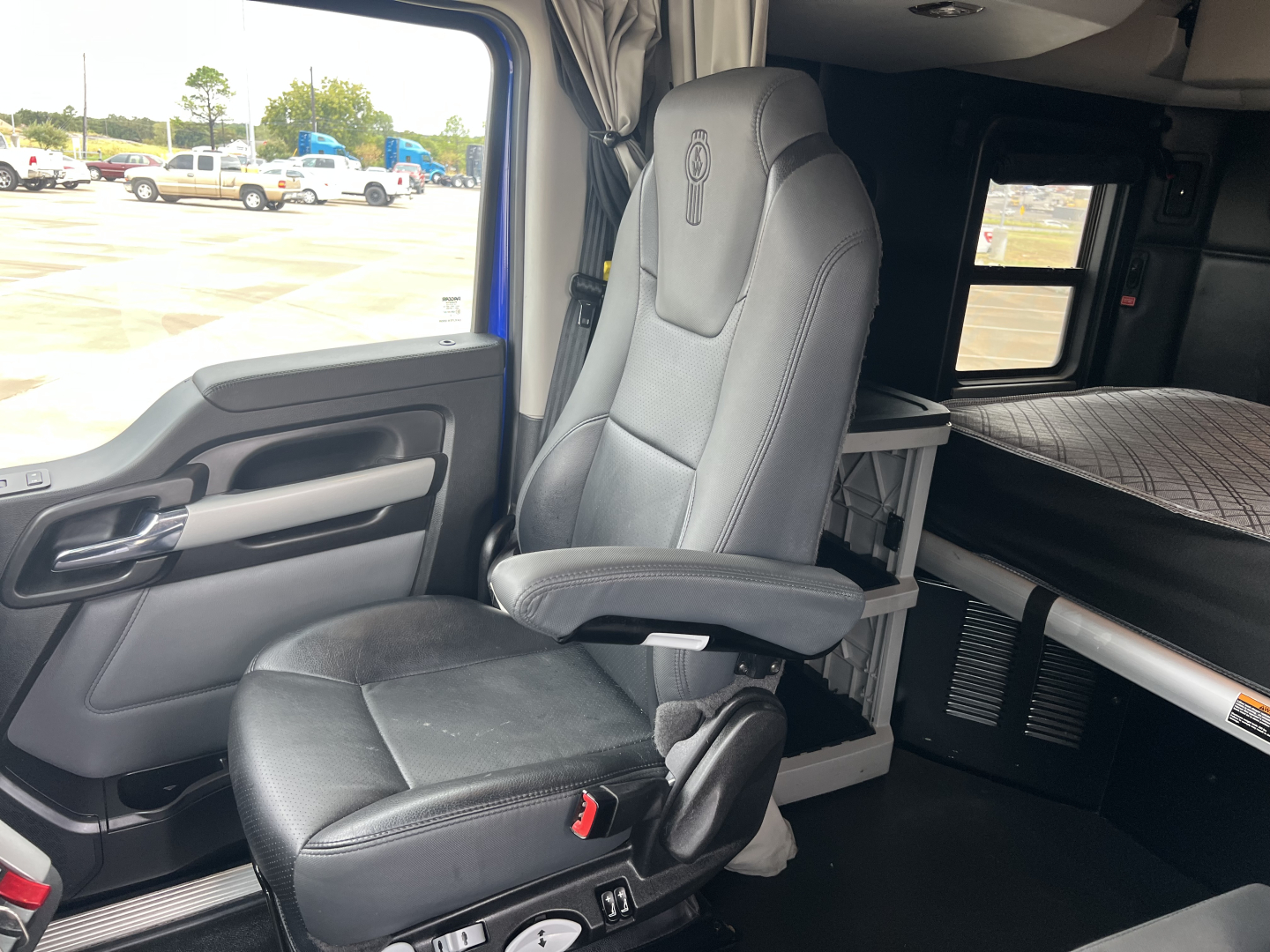 Kenworth T680 cab interior showing driver seat and sleeper area with side door panel and bed in background.