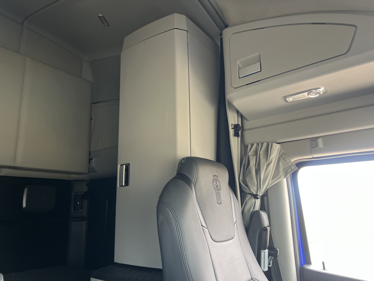Kenworth T680 cabin interior with overhead storage and vertical cabinet, driver seat visible, light gray upholstery, dashboard area, window to the right.