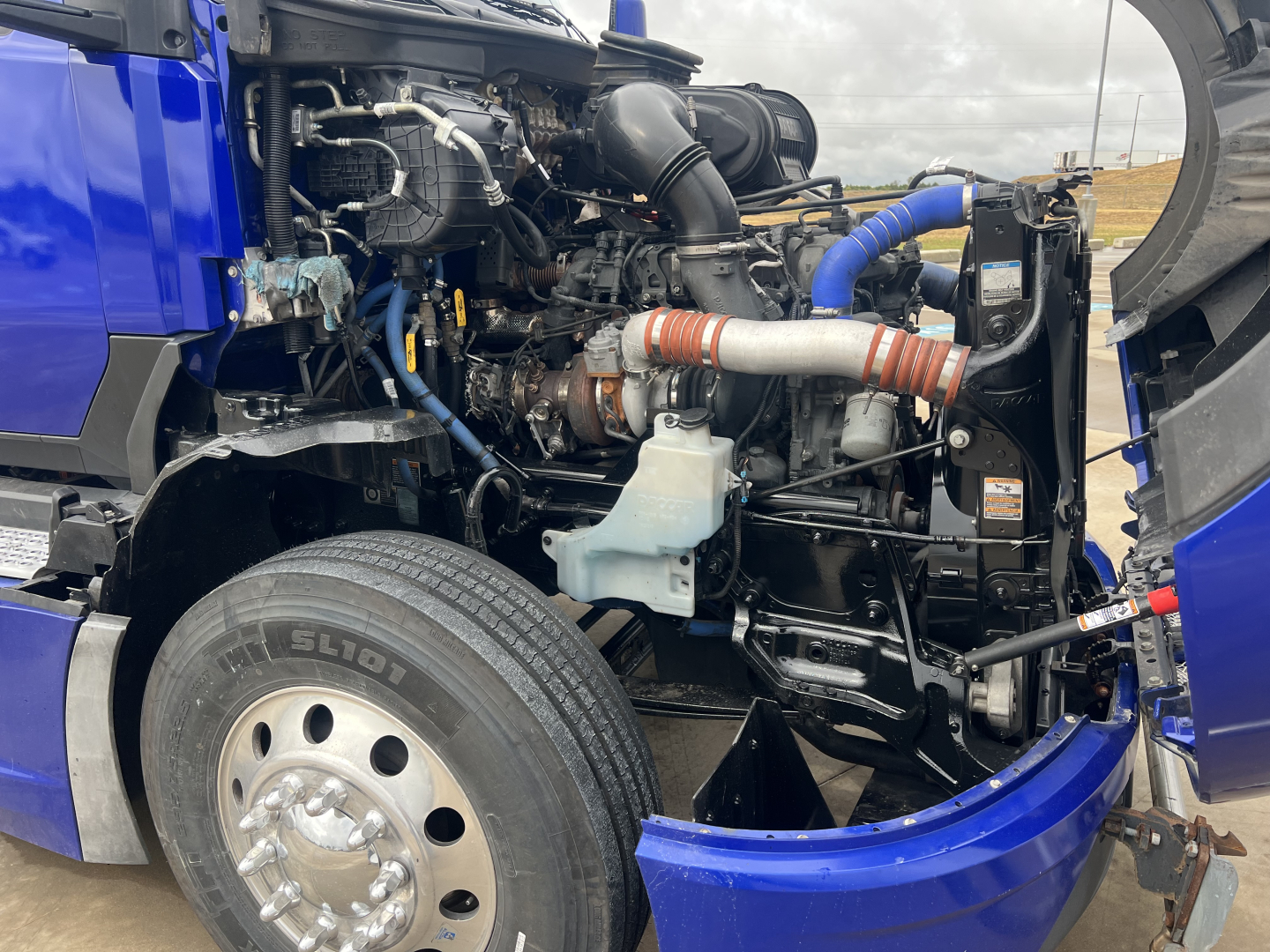 Kenworth T680 engine bay with visible turbo, hoses, and coolant reservoir in blue truck; front wheel and open hood.