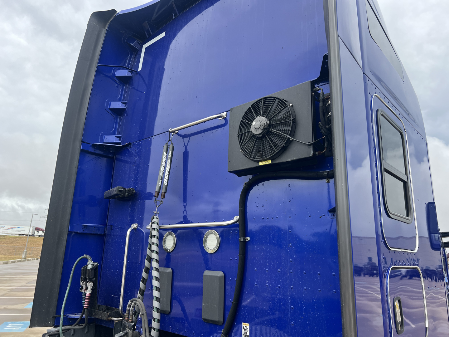 Kenworth T680 sleeper truck rear panel with mounted auxiliary cooling fan and various hoses, gauges, and panels visible on blue exterior.