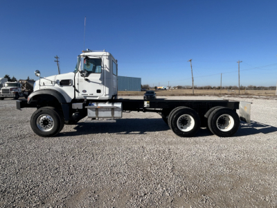 Side view of a white 2026 Mack GR64F cab and chassis truck on a gravel lot with triple rear axles and flatbed.