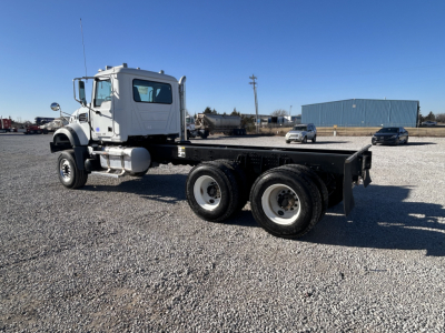 Mack GR64F cab chassis truck in a gravel lot, side view showing chassis and dual rear axles, cab with mirrors and steps, no bodybed installed.
