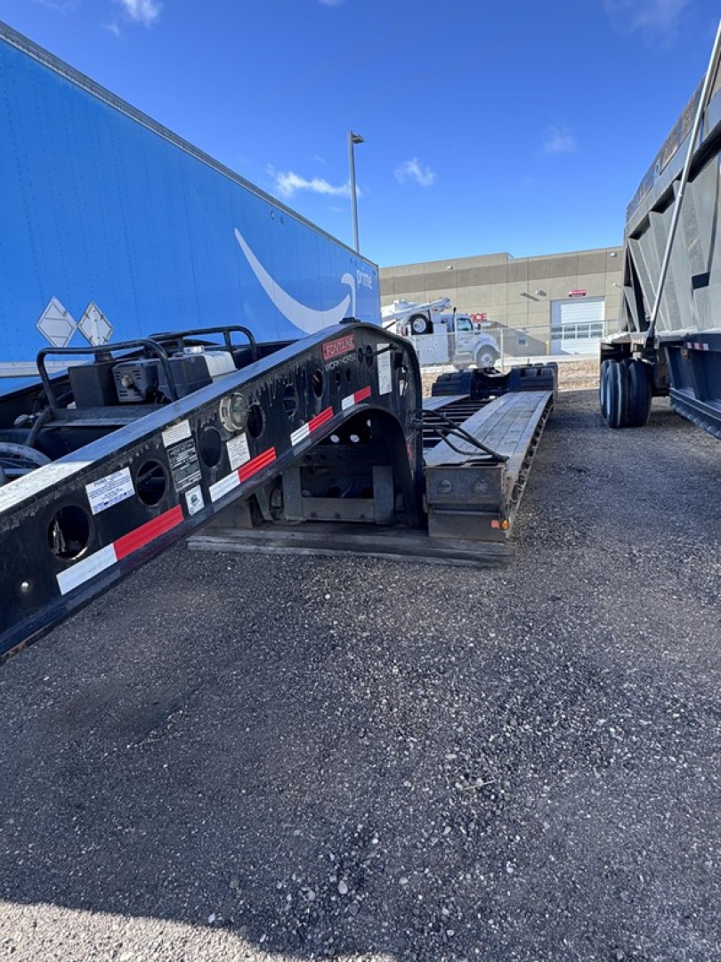 Flatbed trailer in-service showing lowboy deck with pump and wheel wells; blue Fontaine trailer in background; tires and metal frame visible.