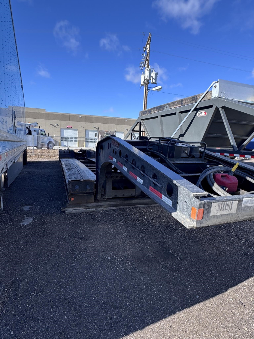 55-foot Fontaine lowboy trailer frame with silver metal, side ramps, and toolboxes. Visible markings include a black frame with diamond plate, rear taillights, and a red battery pack.