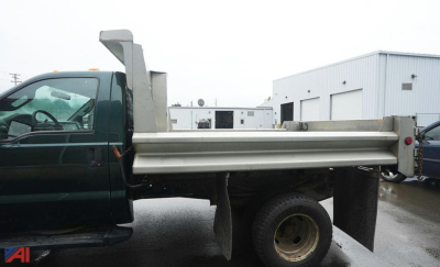 Ford F-350 dump truck side view showing dumping bed and hydraulic lift, green cab with side mirror, gray metal bed, wheel and tire visible.