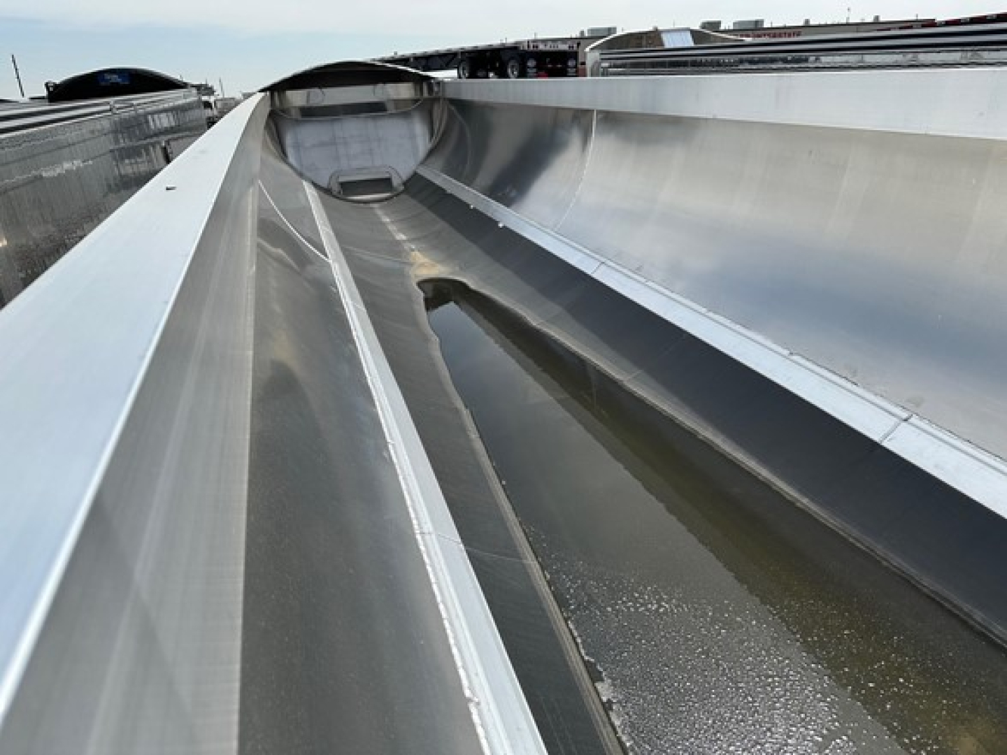 Dump trailer interior showing aluminum side walls and a shallow, water-filled trough along the floor; shiny metallic surfaces, smooth ribs, and a curved corner at the front.