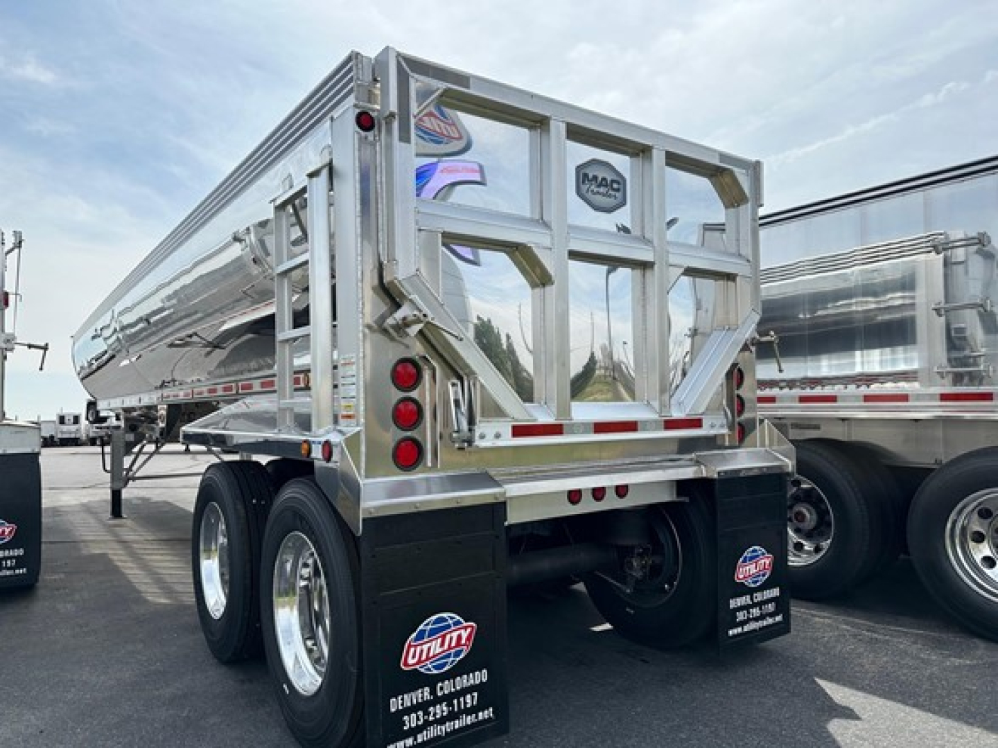 Dump trailer by Mac Simsimer model showing dual rear axles, chrome frame, and rear gate with MAC logo; reflective metal surface and mudflaps with utility logos.