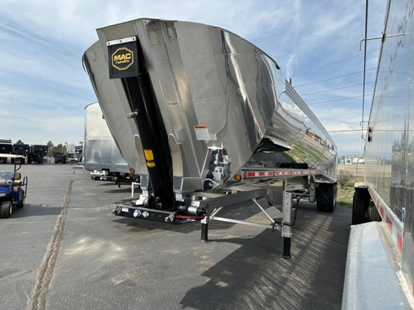 Dump trailer with a MacMacsimiser hopper showing polished aluminum exterior, black hitch, and front support legs; Mac branding on front plate.