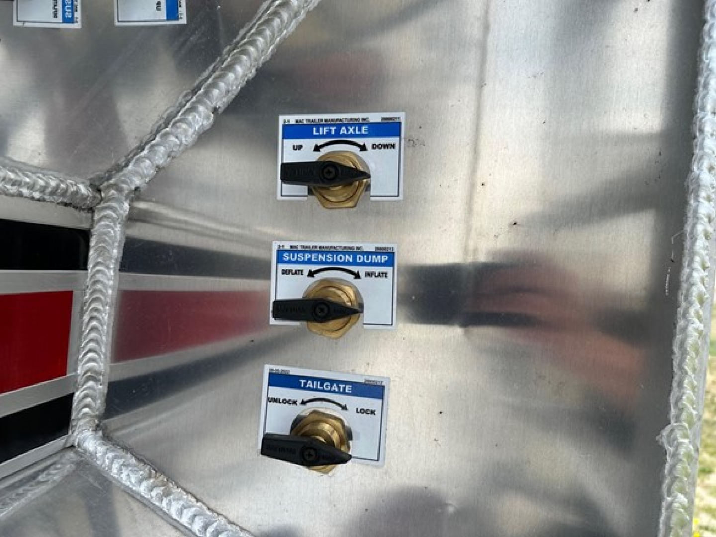Close-up of a metal trailer control panel with three labeled levers: LIFT AXLE UP/DOWN, SUSPENSION DUMP INFLATE/DEF, TAILGATE UNLOCK/LOCK. Blue and white labels on a polished metal surface.