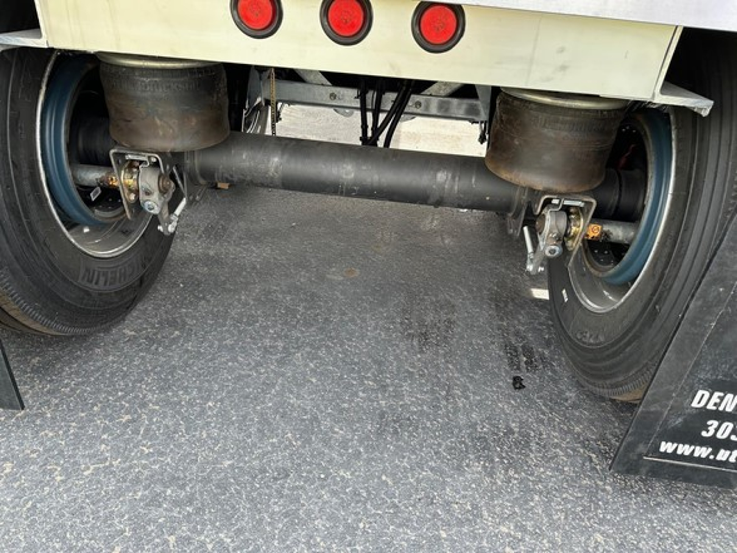 Trailer rear undercarriage showing twin axles with solid drive shaft and suspension, mudflaps, and tires visible.