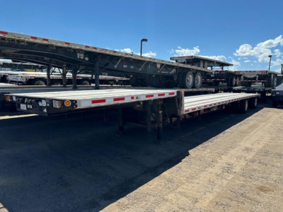 Flatbed trailer with a drop deck in an outdoor lot; visible markings include reflective tape along the side and axle configuration undercarriage. No legible brand or model text visible.